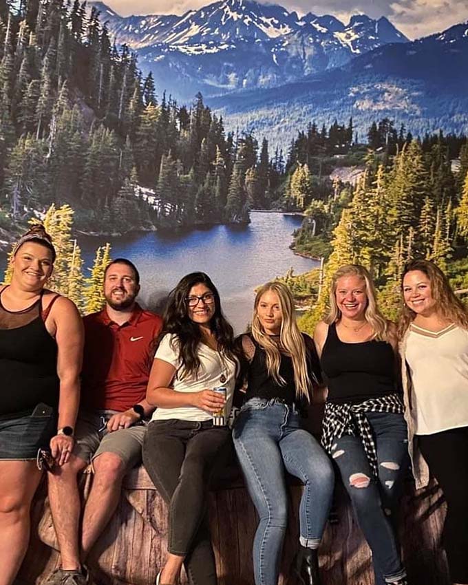 Group of friends sitting on a wood bench, nature painting in background. Montana Nights Top Axe-Bar + Entertainment Center in CT