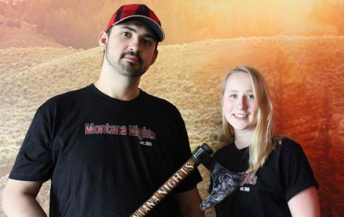 A man and woman stand together, smiling while holding an axe in a forested area. 