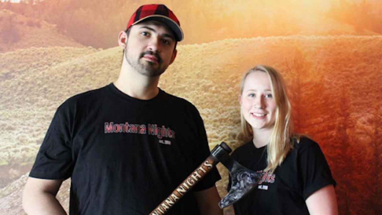 A man and woman stand together, smiling while holding an axe in a forested area. 