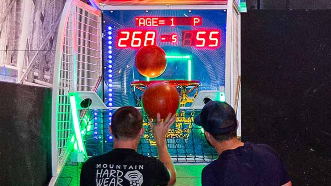 Two people playing an arcade basketball shooting game with a digital scoreboard.