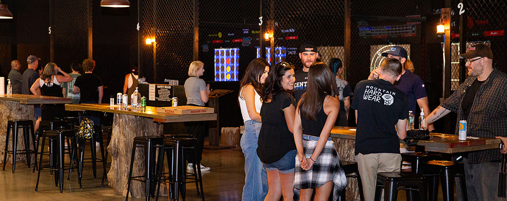 Group of people socializing inside Montana Nights with axe throwing lanes in the background