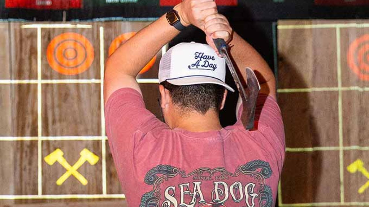 A person at an indoor axe throwing lane, holding an axe overhead and aiming at a wooden target