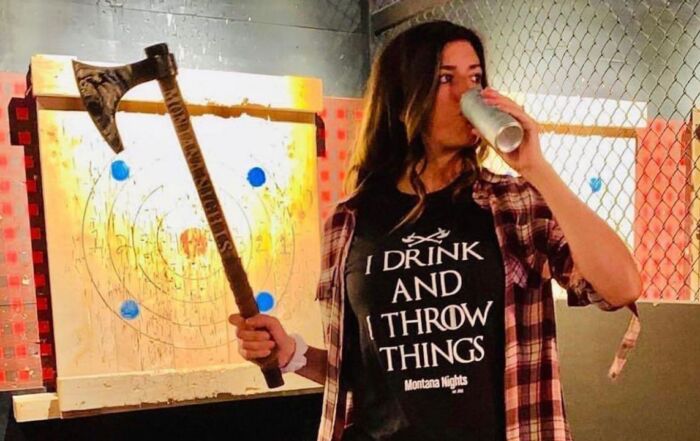 Woman holding an axe and drinking a beverage at a Montana Nights axe throwing lane.