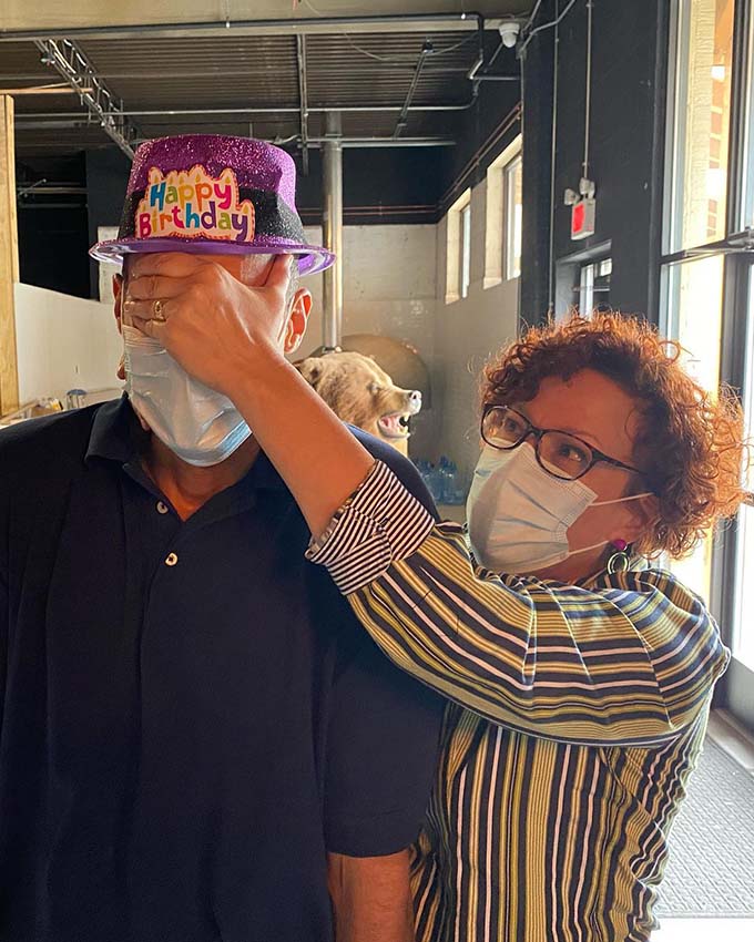 Woman wearing a face mask covering a man's eyes while he wears a purple glittery Happy Birthday hat.